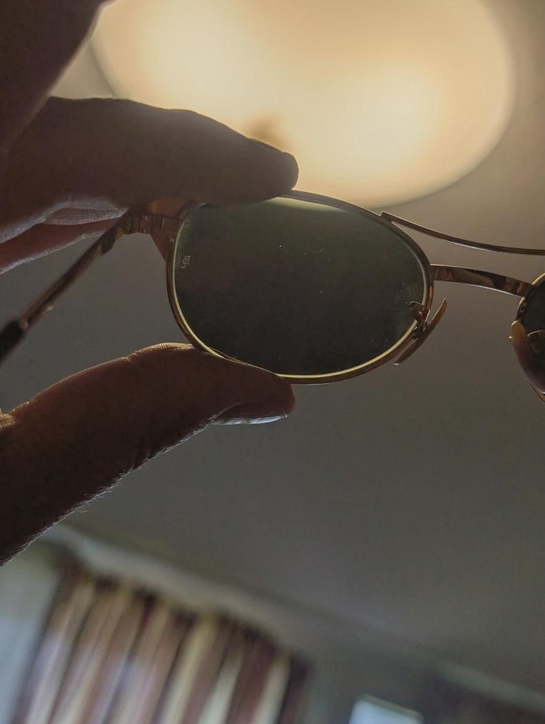 May include: Gold-rimmed sunglasses with dark green lenses. The sunglasses have a classic, round shape and a thin metal frame. The image is taken indoors, with a blurred background and a hand holding the sunglasses.