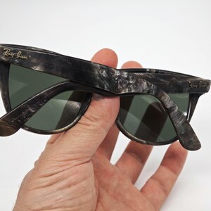Puede incluir: Gafas de sol veteadas con lentes verdes. Las monturas presentan un patr&oacute;n distintivo de remolinos negros, grises y blancos. El logotipo de Ray-Ban es visible en las patillas. Las gafas de sol se sostienen en una mano.