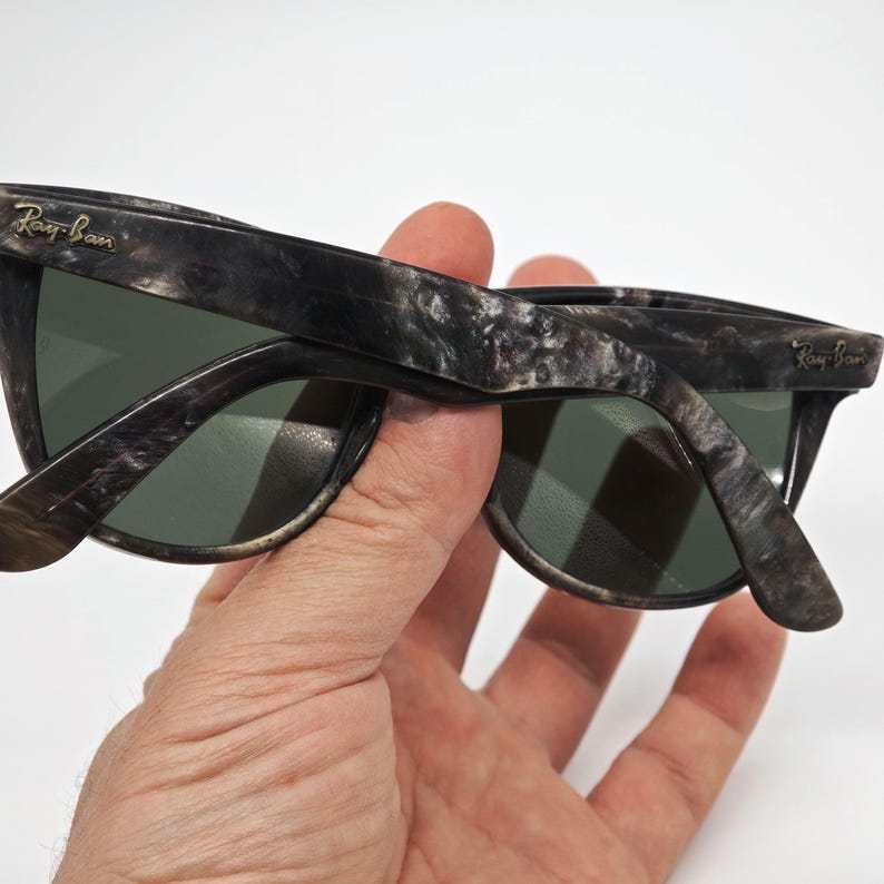 Puede incluir: Gafas de sol veteadas con lentes verdes. Las monturas presentan un patr&oacute;n distintivo de remolinos negros, grises y blancos. El logotipo de Ray-Ban es visible en las patillas. Las gafas de sol se sostienen en una mano.