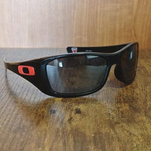 K&ouml;nnte beinhalten: Schwarze Sonnenbrille mit Wrap-around-Design und dunklen Gl&auml;sern. Die schwarzen Rahmen haben ein rotes quadratisches Logo an den B&uuml;geln. Die Sonnenbrille liegt auf einer Holzoberfl&auml;che.