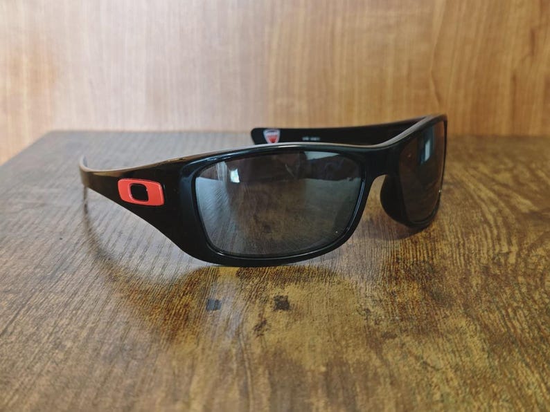 K&ouml;nnte beinhalten: Schwarze Sonnenbrille mit Wrap-around-Design und dunklen Gl&auml;sern. Die schwarzen Rahmen haben ein rotes quadratisches Logo an den B&uuml;geln. Die Sonnenbrille liegt auf einer Holzoberfl&auml;che.