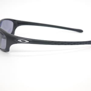 May include: Black sunglasses with a matte finish. The frames have a curved design with a silver logo on the side. The arms have a textured grip for a secure fit. The lenses are a dark gray color.
