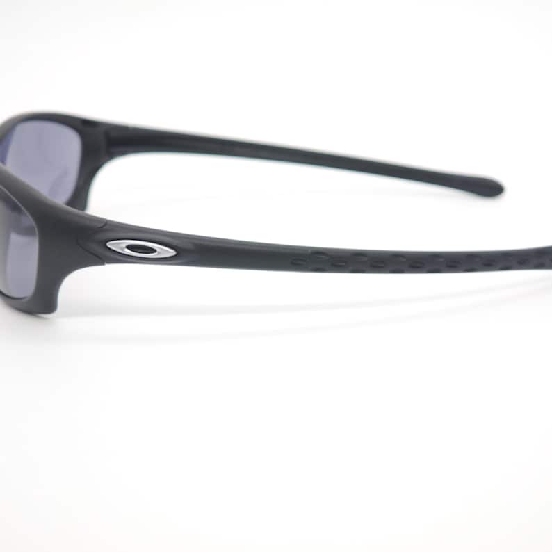 May include: Black sunglasses with a matte finish. The frames have a curved design with a silver logo on the side. The arms have a textured grip for a secure fit. The lenses are a dark gray color.