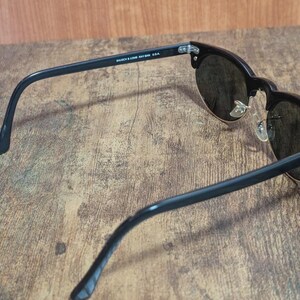 May include: Black sunglasses with dark lenses and a gold-colored rim. The sunglasses have a classic design with a thick black frame and arms. The text "BAUSCH & LOMB RAY-BAN USA" is printed on the arm.
