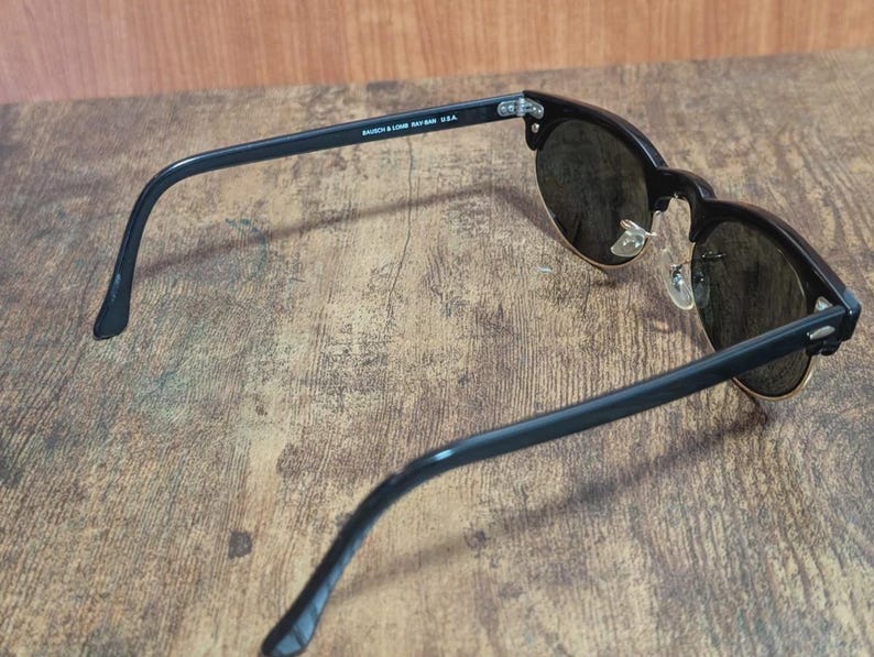 May include: Black sunglasses with dark lenses and a gold-colored rim. The sunglasses have a classic design with a thick black frame and arms. The text "BAUSCH & LOMB RAY-BAN USA" is printed on the arm.