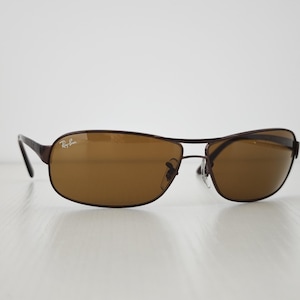 May include: Dark brown metal frame sunglasses with a rectangular shape and brown tinted lenses. The Ray-Ban logo is printed in white on the upper corner of the left lens. The eyewear features thin metal temples and adjustable nose pads.