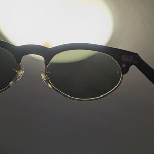 May include: Black and gold-rimmed sunglasses with dark lenses. The sunglasses have a classic design with a black upper frame and gold-colored lower rims. The lenses are dark, providing shade and protection from the sun.