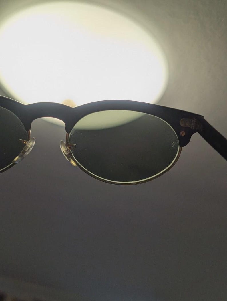 May include: Black and gold-rimmed sunglasses with dark lenses. The sunglasses have a classic design with a black upper frame and gold-colored lower rims. The lenses are dark, providing shade and protection from the sun.