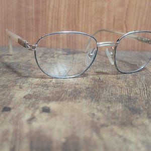 Può includere: Occhiali con montatura argentata di forma ovale classica. Le montature sottili e delicate incorniciano lenti trasparenti. Le aste sono beige chiaro. Gli occhiali sono appoggiati su uno sfondo di legno.