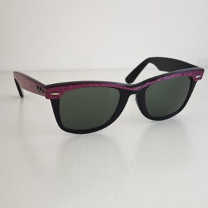 Puede incluir: Gafas de sol Ray-Ban con montura negra y un patr&oacute;n de veta de madera p&uacute;rpura texturizada a lo largo de la parte superior y las patillas. Las lentes son de color verde oscuro y las patillas incluyen detalles met&aacute;licos plateados con el logo.