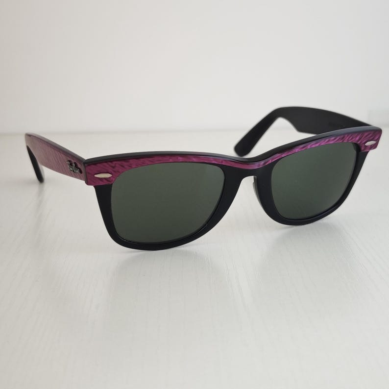 Puede incluir: Gafas de sol Ray-Ban con montura negra y un patr&oacute;n de veta de madera p&uacute;rpura texturizada a lo largo de la parte superior y las patillas. Las lentes son de color verde oscuro y las patillas incluyen detalles met&aacute;licos plateados con el logo.
