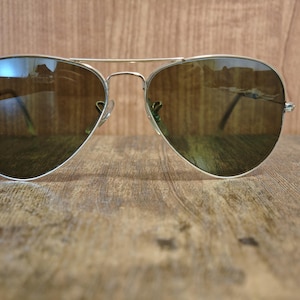May include: Aviator sunglasses with a silver frame and brown lenses. The Ray-Ban logo is visible on the upper left lens. The sunglasses are resting on a wooden surface.