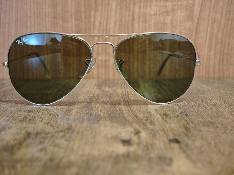 May include: Aviator sunglasses with a silver frame and brown lenses. The Ray-Ban logo is visible on the upper left lens. The sunglasses are resting on a wooden surface.
