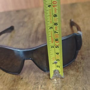 May include: Black sunglasses with dark lenses are measured with a yellow tape measure. The sunglasses are approximately 2 inches (5 cm) tall. The tape measure shows the measurement in inches and centimeters.