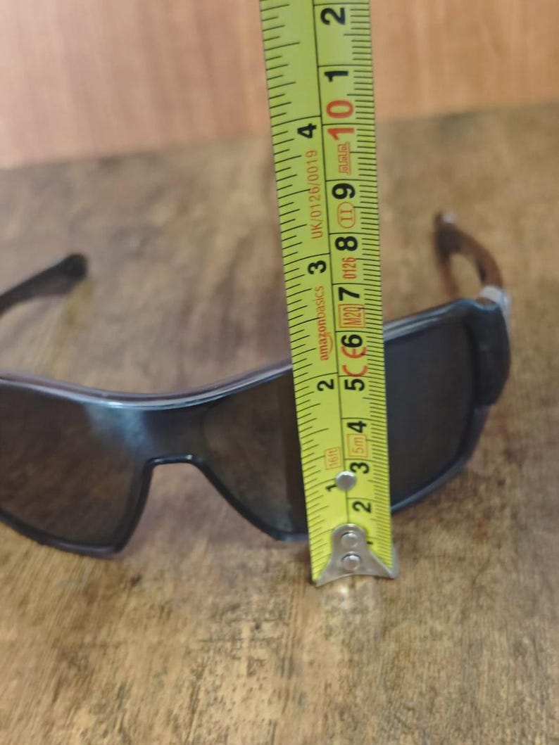 May include: Black sunglasses with dark lenses are measured with a yellow tape measure. The sunglasses are approximately 2 inches (5 cm) tall. The tape measure shows the measurement in inches and centimeters.