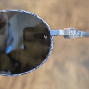 May include: Oval-shaped sunglasses with a silver frame and dark lenses. The frame has a subtle pattern with blue and white accents. The temples feature a decorative design and the initials "BL".