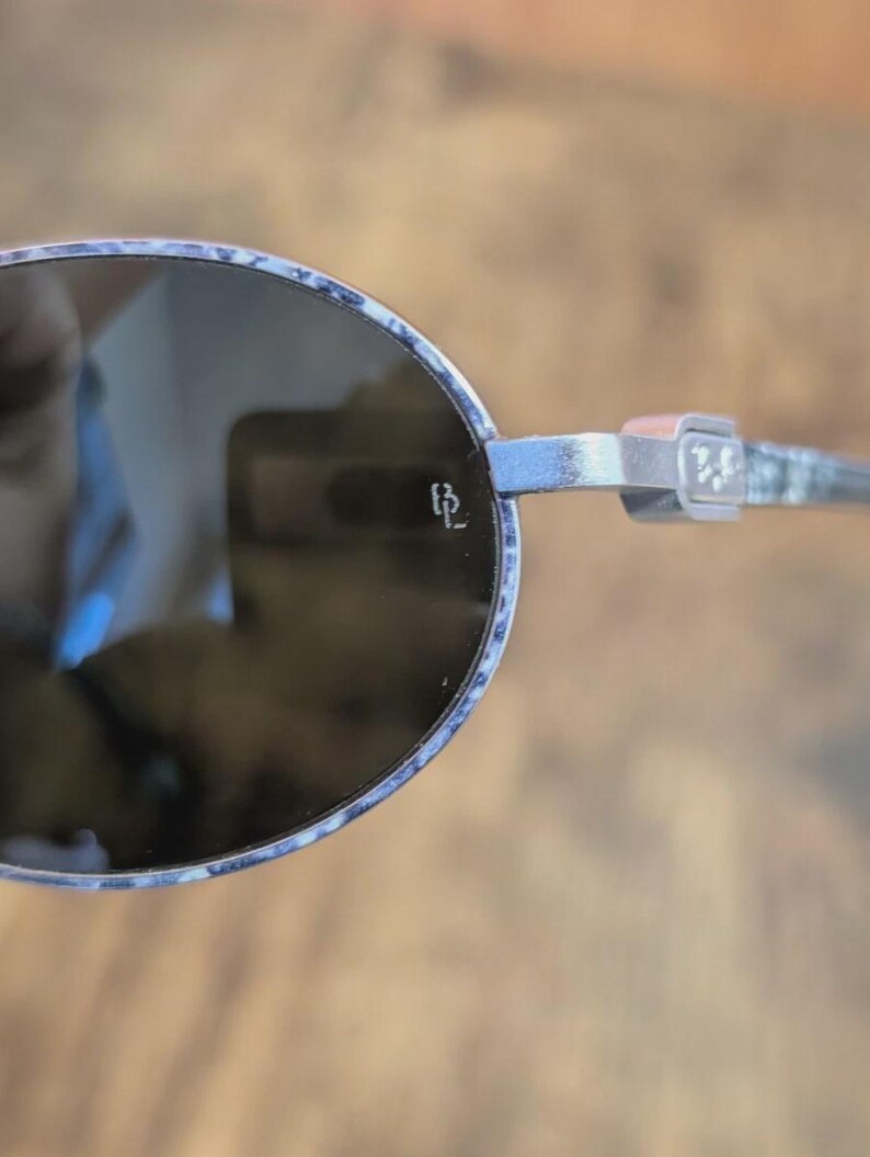 May include: Oval-shaped sunglasses with a silver frame and dark lenses. The frame has a subtle pattern with blue and white accents. The temples feature a decorative design and the initials "BL".