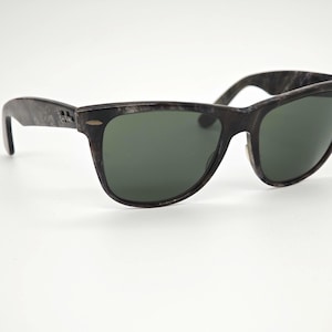 Puede incluir: Gafas de sol gris oscuro con un patr&oacute;n veteado en las monturas y lentes verde oscuro. Las gafas de sol tienen una forma rectangular cl&aacute;sica y un peque&ntilde;o logotipo en la patilla.