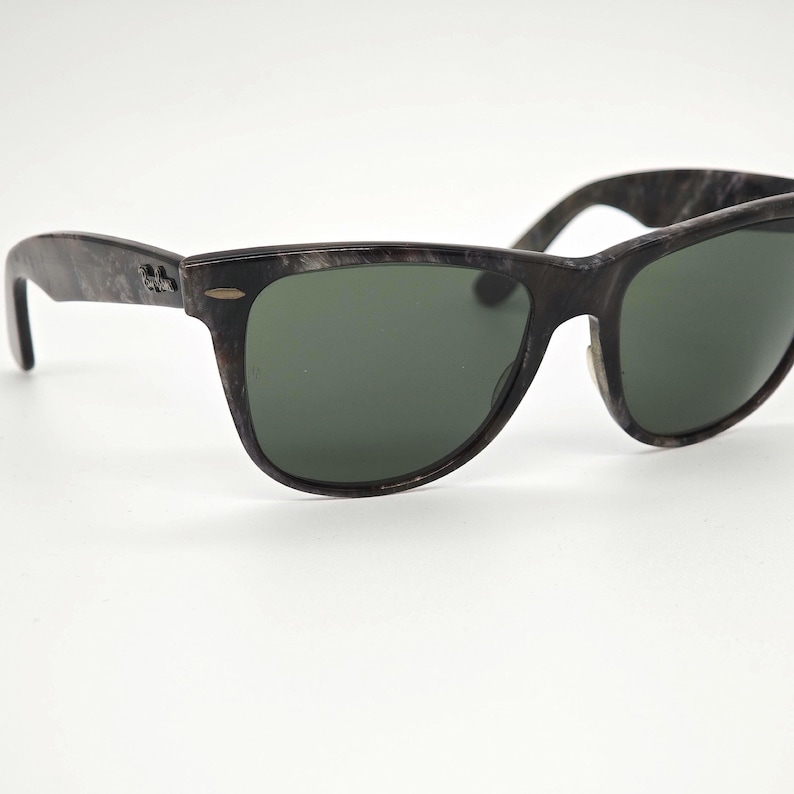 Puede incluir: Gafas de sol gris oscuro con un patr&oacute;n veteado en las monturas y lentes verde oscuro. Las gafas de sol tienen una forma rectangular cl&aacute;sica y un peque&ntilde;o logotipo en la patilla.