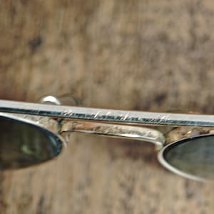 May include: Silver-framed sunglasses with oval lenses. The metal frame shows signs of wear. The lenses are a dark color, providing shade. The sunglasses are set against a blurred, brown background.