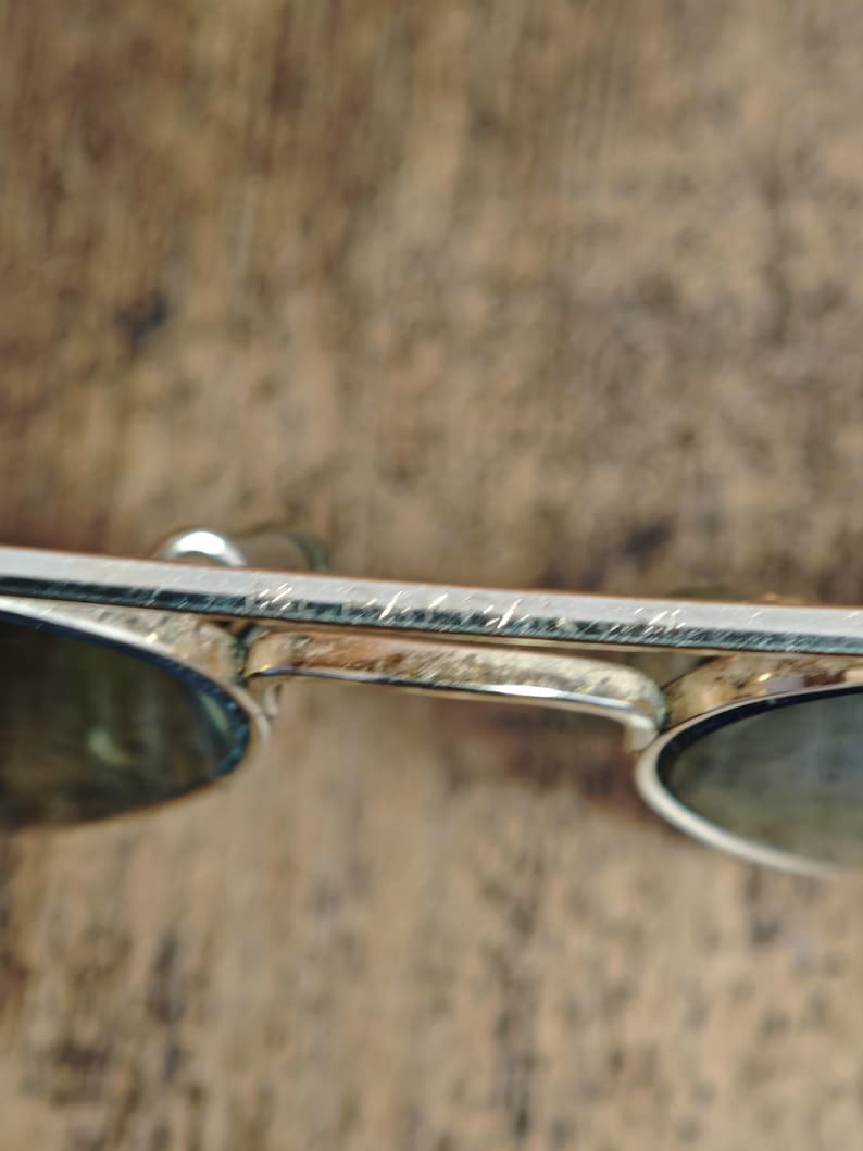 May include: Silver-framed sunglasses with oval lenses. The metal frame shows signs of wear. The lenses are a dark color, providing shade. The sunglasses are set against a blurred, brown background.