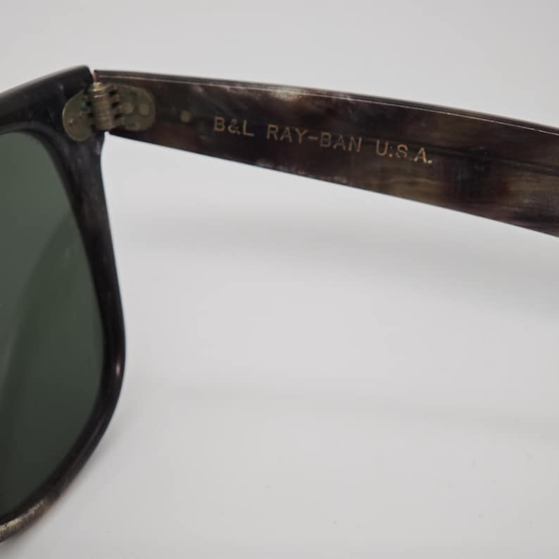 Puede incluir: Primer plano de unas gafas de sol con montura oscura y lentes verdes. El texto "B&L RAY-BAN U.S.A." est&aacute; impreso en la patilla. La montura tiene un patr&oacute;n moteado.