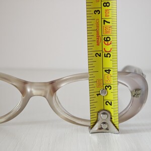 May include: Translucent beige oval-shaped eyeglasses frames positioned next to a yellow measuring tape. The tape shows the lens height is approximately 1.5 inches. The frames feature a smooth, rounded design with a small metallic accent on the corner.