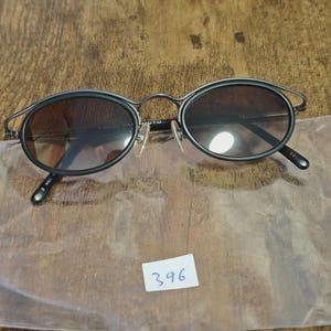 May include: Black oval sunglasses with a thin metal frame and tinted lenses. The sunglasses have a unique design with a curved top bar. The lenses are a gradient brown color. The sunglasses are on a wooden surface.