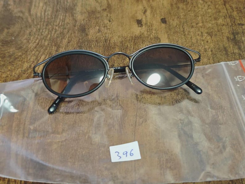 May include: Black oval sunglasses with a thin metal frame and tinted lenses. The sunglasses have a unique design with a curved top bar. The lenses are a gradient brown color. The sunglasses are on a wooden surface.