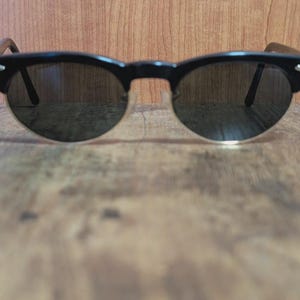 May include: Black sunglasses with a semi-rimless design. The frames are black with a rounded shape, and the lenses are dark. The arms of the sunglasses are a dark brown color. The sunglasses are resting on a wooden surface.