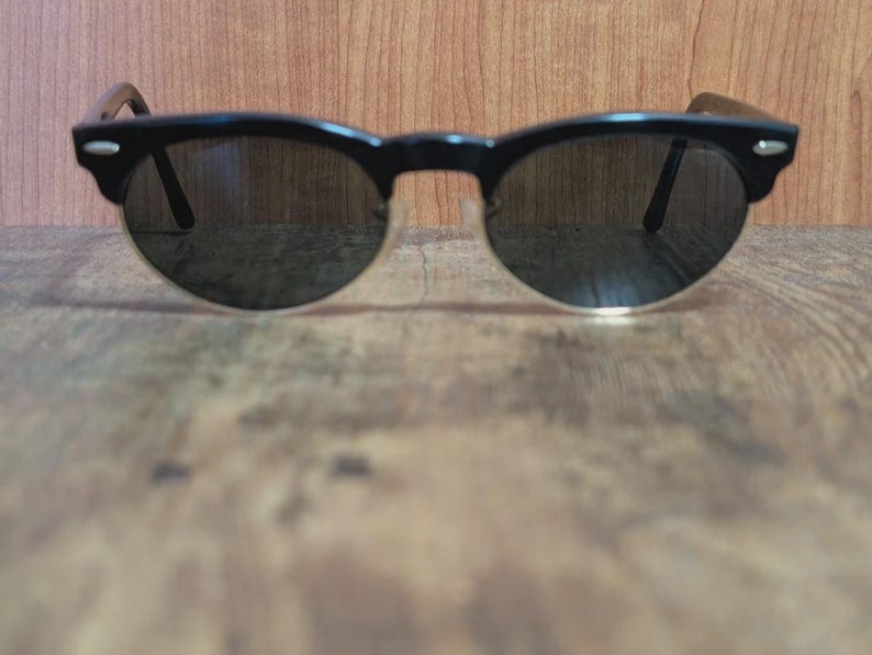 May include: Black sunglasses with a semi-rimless design. The frames are black with a rounded shape, and the lenses are dark. The arms of the sunglasses are a dark brown color. The sunglasses are resting on a wooden surface.