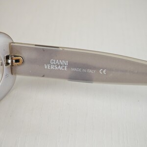 May include: Translucent light gray eyeglasses temple arm featuring the text GIANNI VERSACE MADE IN ITALY and a CE mark printed in white on the inner side. The frame includes a metallic hinge component connecting the arm to the front piece.