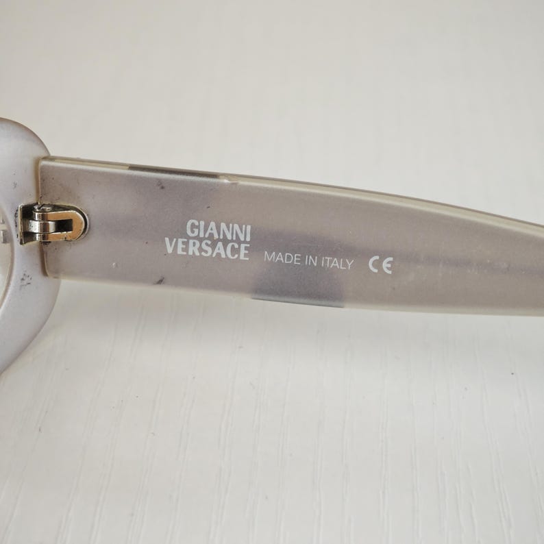 May include: Translucent light gray eyeglasses temple arm featuring the text GIANNI VERSACE MADE IN ITALY and a CE mark printed in white on the inner side. The frame includes a metallic hinge component connecting the arm to the front piece.