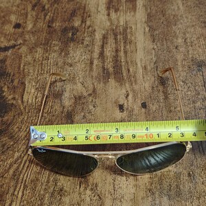 May include: Gold-framed sunglasses with dark lenses, measuring approximately 5 inches in width. The Ray-Ban logo is visible on the left lens. The glasses are set against a wooden background.