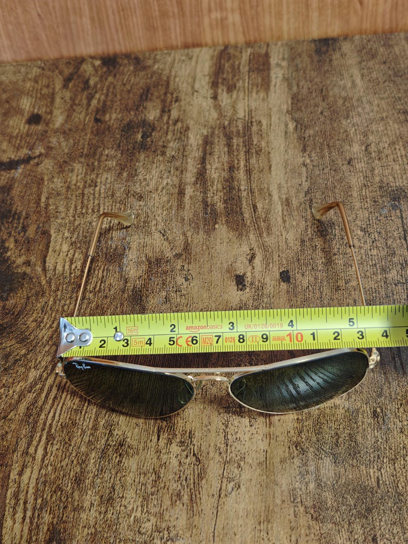 May include: Gold-framed sunglasses with dark lenses, measuring approximately 5 inches in width. The Ray-Ban logo is visible on the left lens. The glasses are set against a wooden background.