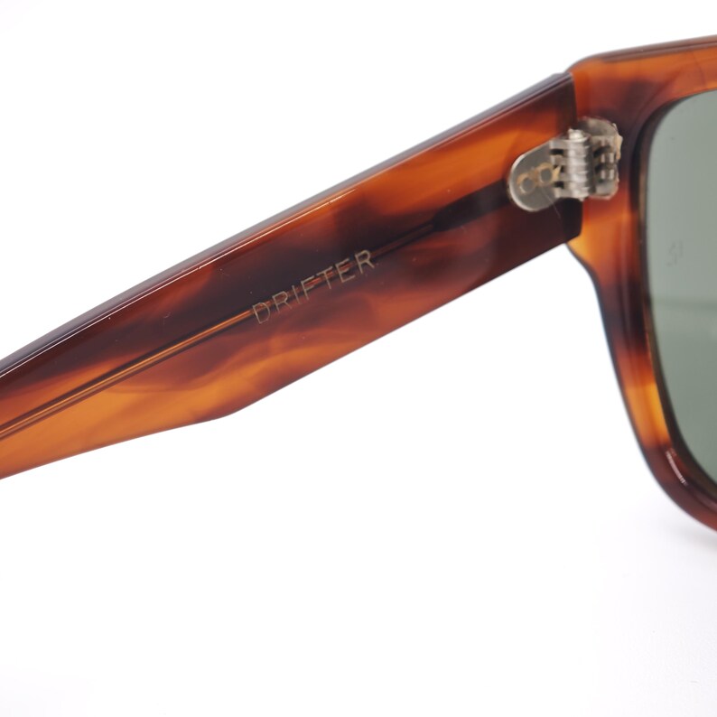 May include: Close-up of sunglasses with a tortoiseshell pattern frame. The word "DRIFTER" is printed on the arm. The lenses are a muted green color. The hinge is silver.