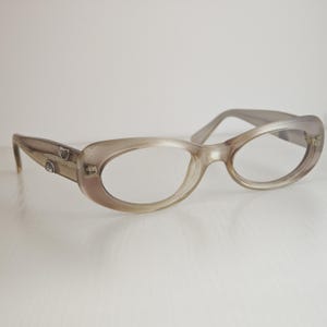 May include: Oval-shaped eyeglasses with a translucent, light taupe-colored frame. The temples feature small, silver-toned metallic accents embedded near the hinges, providing a subtle contrast to the smooth, rounded plastic construction of the eyewear.