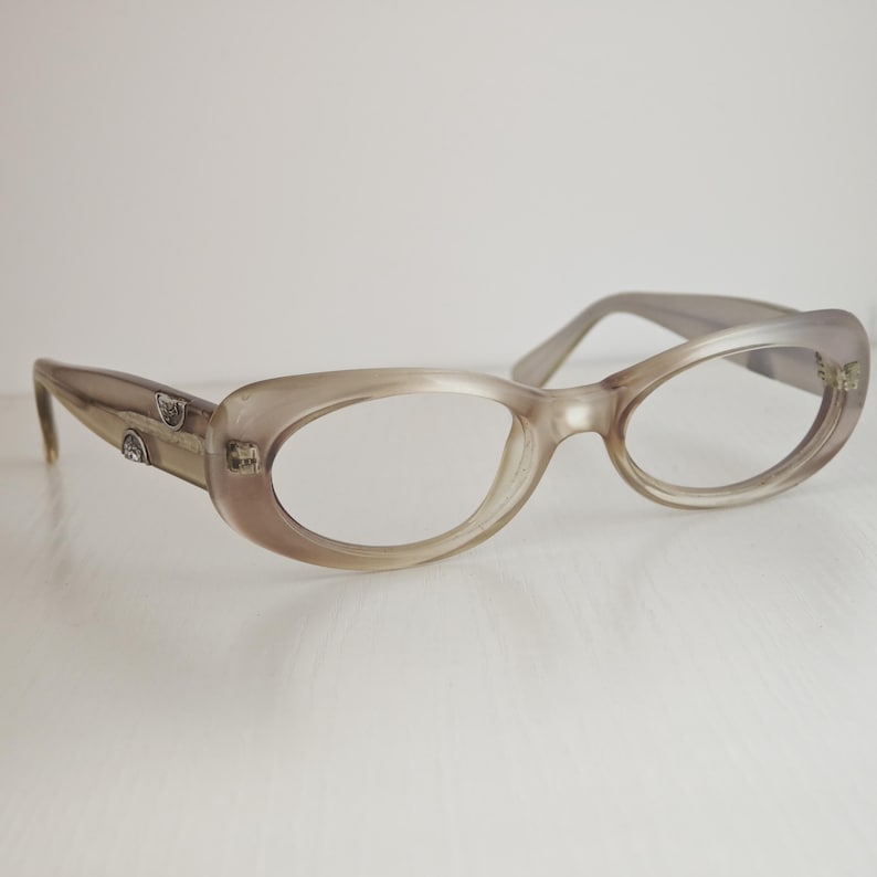 May include: Oval-shaped eyeglasses with a translucent, light taupe-colored frame. The temples feature small, silver-toned metallic accents embedded near the hinges, providing a subtle contrast to the smooth, rounded plastic construction of the eyewear.