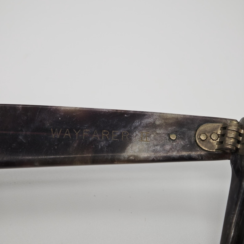 Puede incluir: Primer plano de gafas de sol estilo Wayfarer. La montura oscura y moteada lleva la palabra "WAYFARER" en letras doradas. La bisagra y la patilla son visibles.
