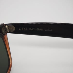 May include: Close-up of a pair of Ray-Ban sunglasses. The frame is black with a brown-orange accent. The text "B&L RAY-BAN U.S.A." is printed on the arm. The lenses are a dark green color.
