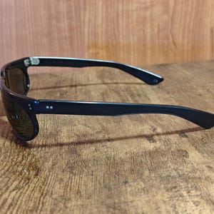 May include: Black sunglasses with dark lenses. The sunglasses have a rectangular frame and a sleek, modern design. The arms of the sunglasses are black and have two small, white dots near the hinges. The sunglasses are resting on a wooden surface.