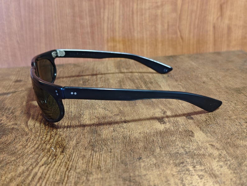 May include: Black sunglasses with dark lenses. The sunglasses have a rectangular frame and a sleek, modern design. The arms of the sunglasses are black and have two small, white dots near the hinges. The sunglasses are resting on a wooden surface.