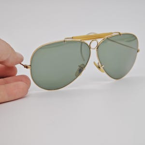 May include: Gold-framed aviator sunglasses with green lenses. The sunglasses have a double bridge and a light-colored bar across the top. The lenses are teardrop-shaped. The sunglasses are held by a hand.