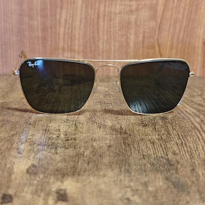 Puede incluir: Gafas de sol con montura dorada y lentes oscuras. Las gafas de sol tienen un dise&ntilde;o cl&aacute;sico de aviador con doble puente y un logotipo sutil en la lente superior izquierda. La montura es dorada brillante y las lentes son oscuras.