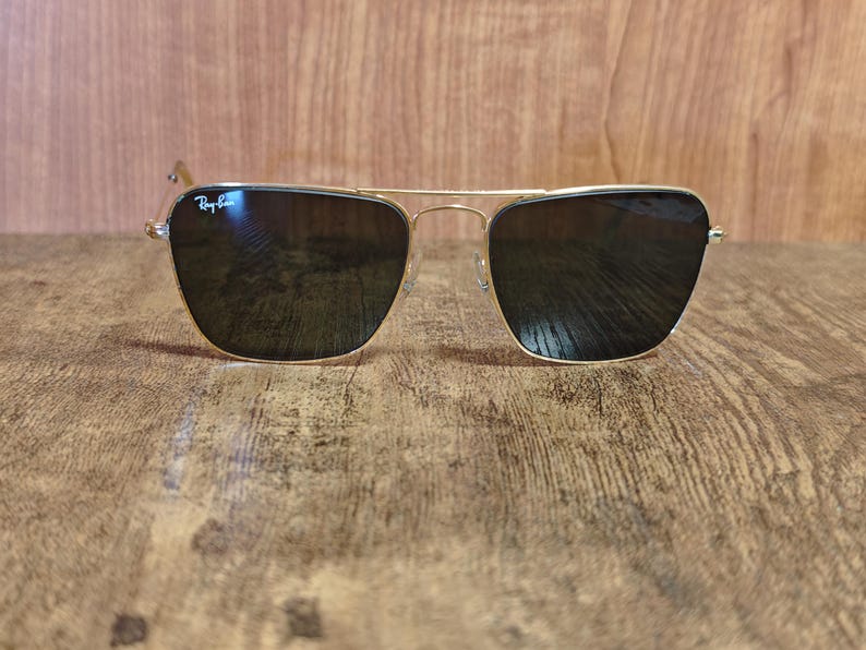 Puede incluir: Gafas de sol con montura dorada y lentes oscuras. Las gafas de sol tienen un dise&ntilde;o cl&aacute;sico de aviador con doble puente y un logotipo sutil en la lente superior izquierda. La montura es dorada brillante y las lentes son oscuras.