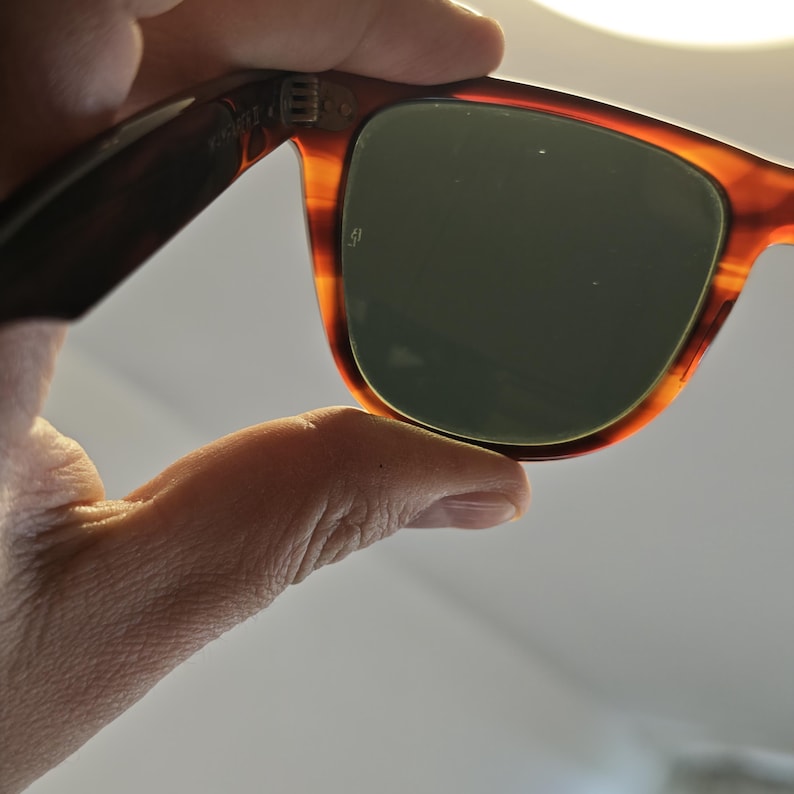 May include: Classic tortoiseshell sunglasses with dark green lenses. The frames have a warm, brown and orange pattern. The sunglasses are held up by a hand, showcasing the design and shape of the eyewear.