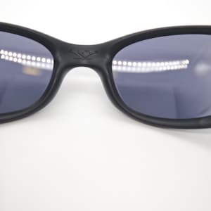 May include: Close-up of black sunglasses with dark lenses. The sunglasses have a curved frame and a small emblem on the bridge. The lenses reflect a bright light source.