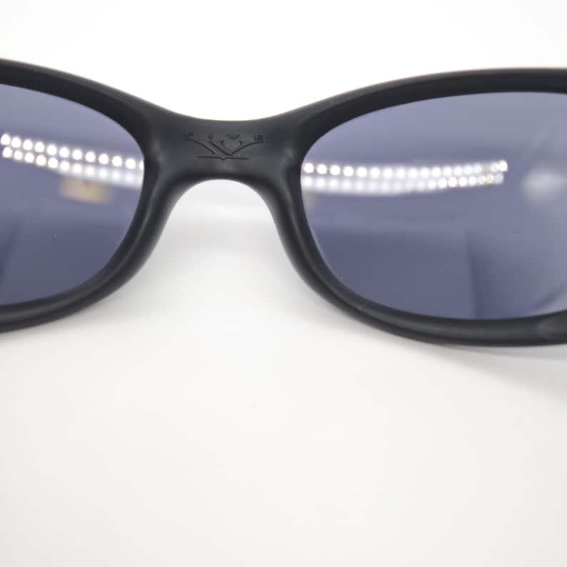 May include: Close-up of black sunglasses with dark lenses. The sunglasses have a curved frame and a small emblem on the bridge. The lenses reflect a bright light source.