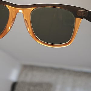 May include: Close-up of sunglasses with a brown frame and amber-colored accents. The lenses are a dark green color. The sunglasses have a classic, retro design.