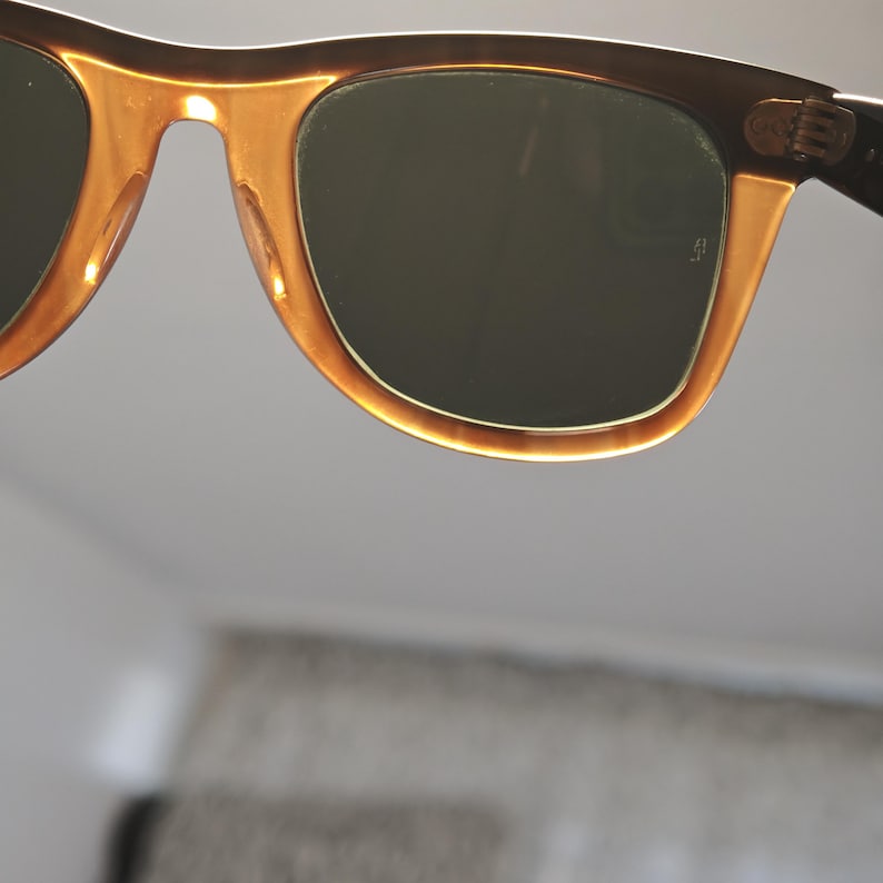 May include: Close-up of sunglasses with a brown frame and amber-colored accents. The lenses are a dark green color. The sunglasses have a classic, retro design.